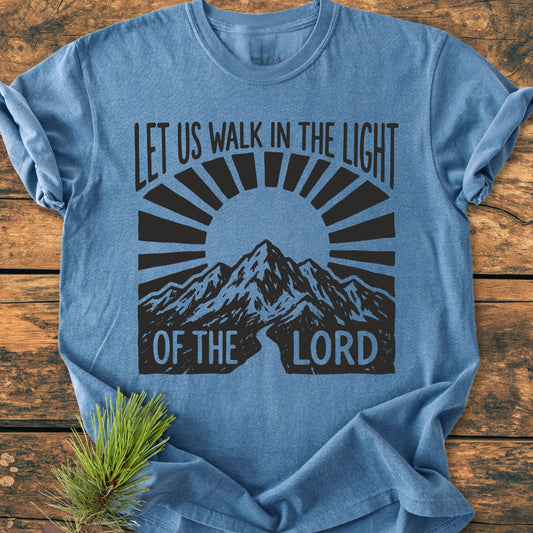 Heather-blue Let Us Walk In The Light Of The Lord - Vintage Wash Tee featuring a mountain range and rising-sun graphic, laid flat on wood, accented by a pine sprig.