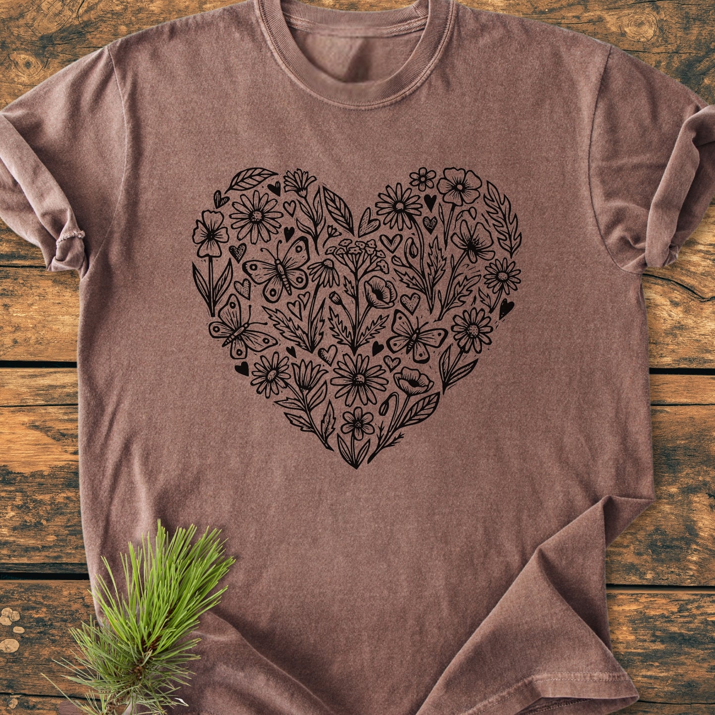 Wildflower Heart Butterfly Soul - Vintage Heavyweight Tee in heather-brown, featuring a black floral heart design and butterflies on the chest, laid flat on a wooden surface with a pine sprig nearby.