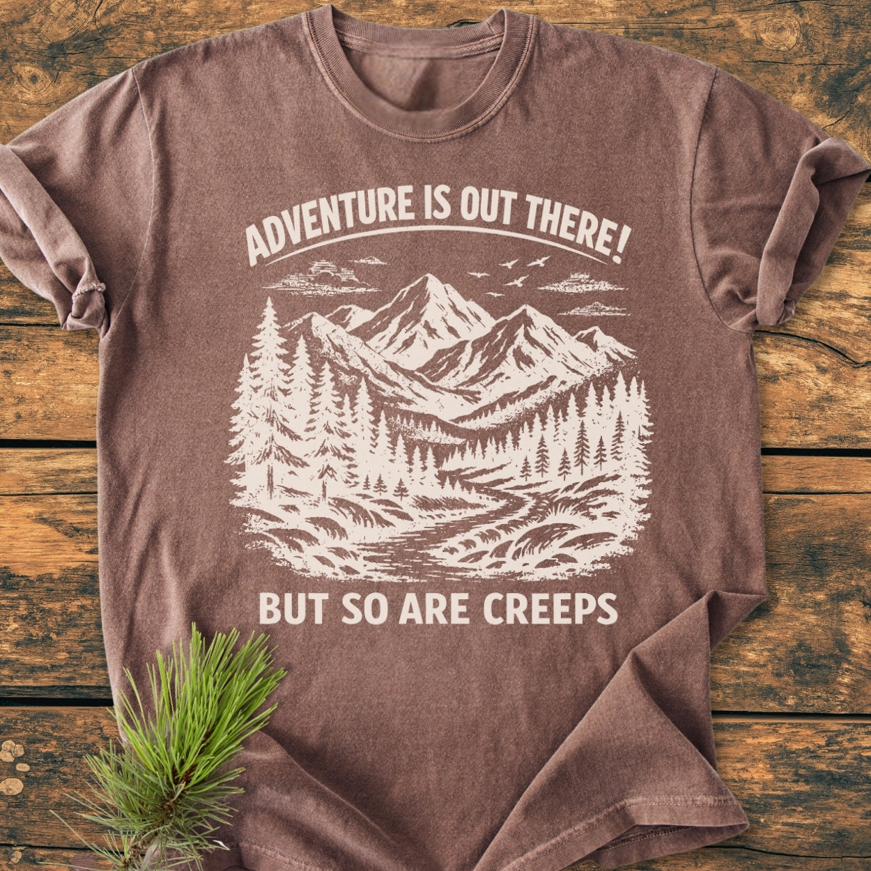 Adventure Is Out There - Vintage Wash Tee features a mountain and pine illustration with playful text, laid on wood. This relaxed, soft-washed, durable tee is perfect for daily wear.