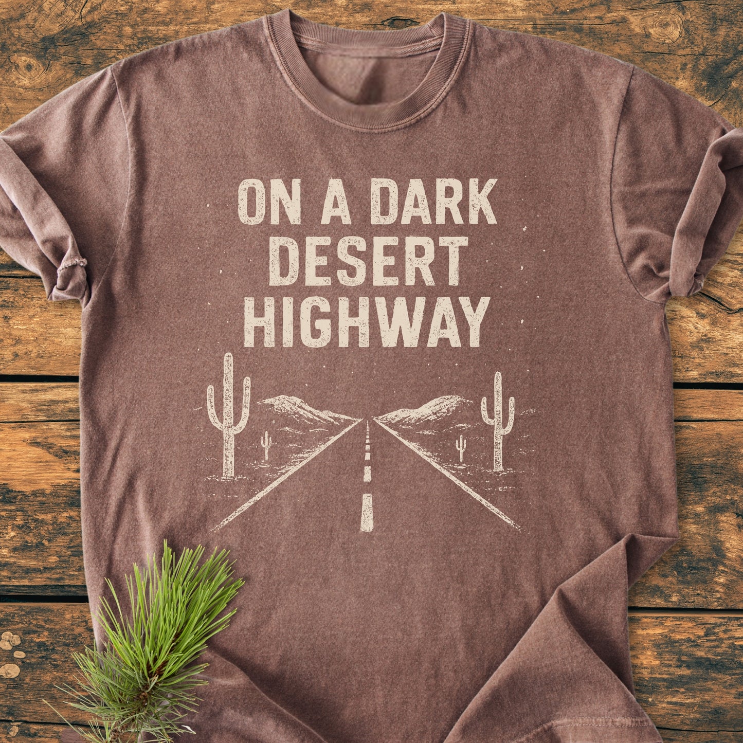 On A Dark Desert Highway - Vintage Wash Tee: Brown short-sleeve shirt with off-white distressed text and desert road illustration, laid flat on wood, complemented by a pine branch for natural touch.