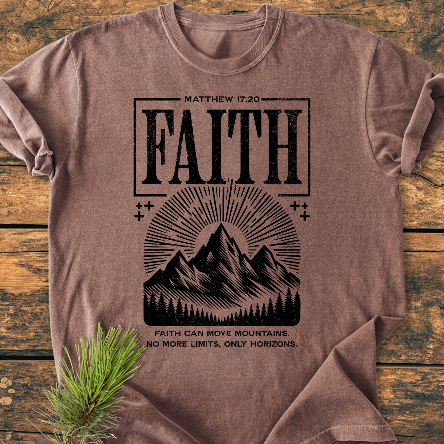 Faith Moves Mountains - Vintage Wash Tee showcasing FAITH and a mountain range with sun-rays. Laid on wood with a pine branch, emphasizing the garment-dyed, soft-washed, relaxed fit, 100% cotton design.