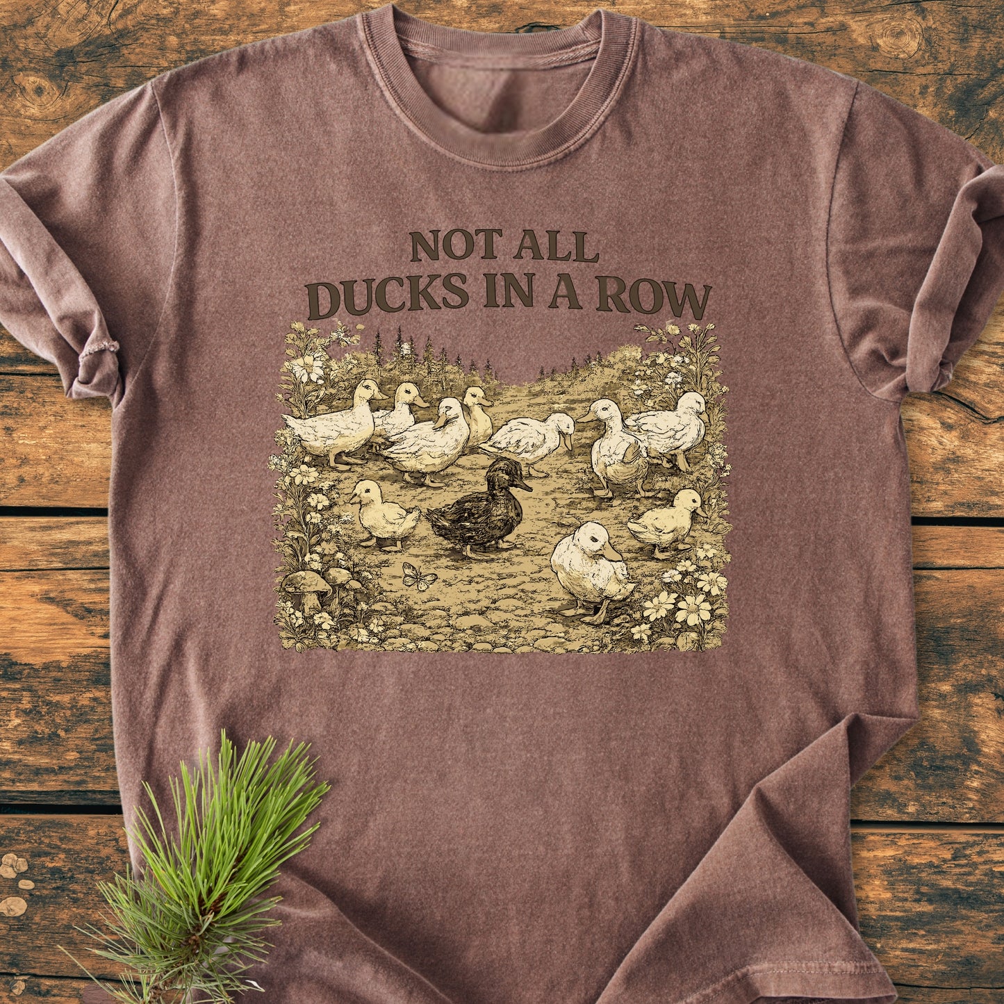 Not All Ducks In A Row - Vintage Wash Tee: Brown T-shirt featuring a sketched duck scene with slogan, laid flat on wood, showcasing its relaxed fit and durable design.