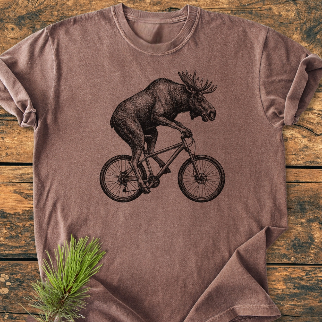 Moose On The Road - Vintage Wash Tee featuring a moose on a bicycle in black line-art on a brown-heather shirt, styled with a pine bough, laid on a rustic wood surface.