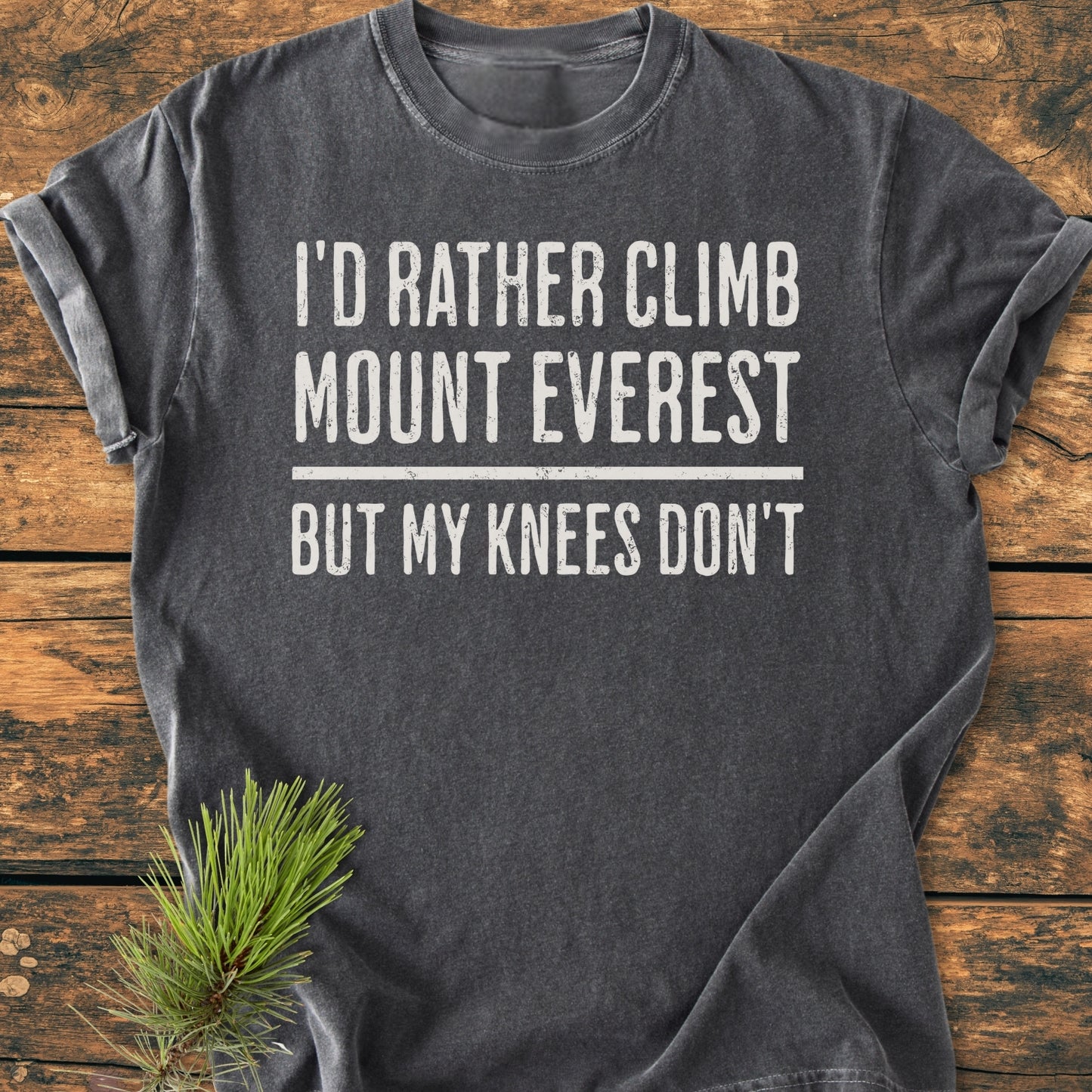 Dark-gray I'd Rather Climb Mount Everest - Vintage Wash Tee featuring a white, distressed slogan, laid on a wooden surface with a pine sprig. Crew-neck, relaxed fit, showcasing soft, garment-dyed fabric.