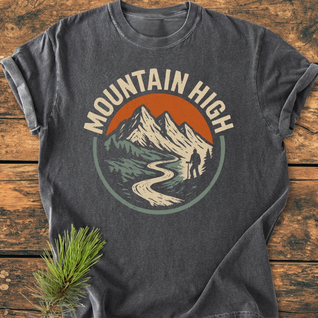 Mountain High - Vintage Wash Tee featuring a circular graphic with snowy peaks, a hiker, and MOUNTAIN HIGH text, on a dark-gray, short-sleeve T-shirt laid flat on wood.