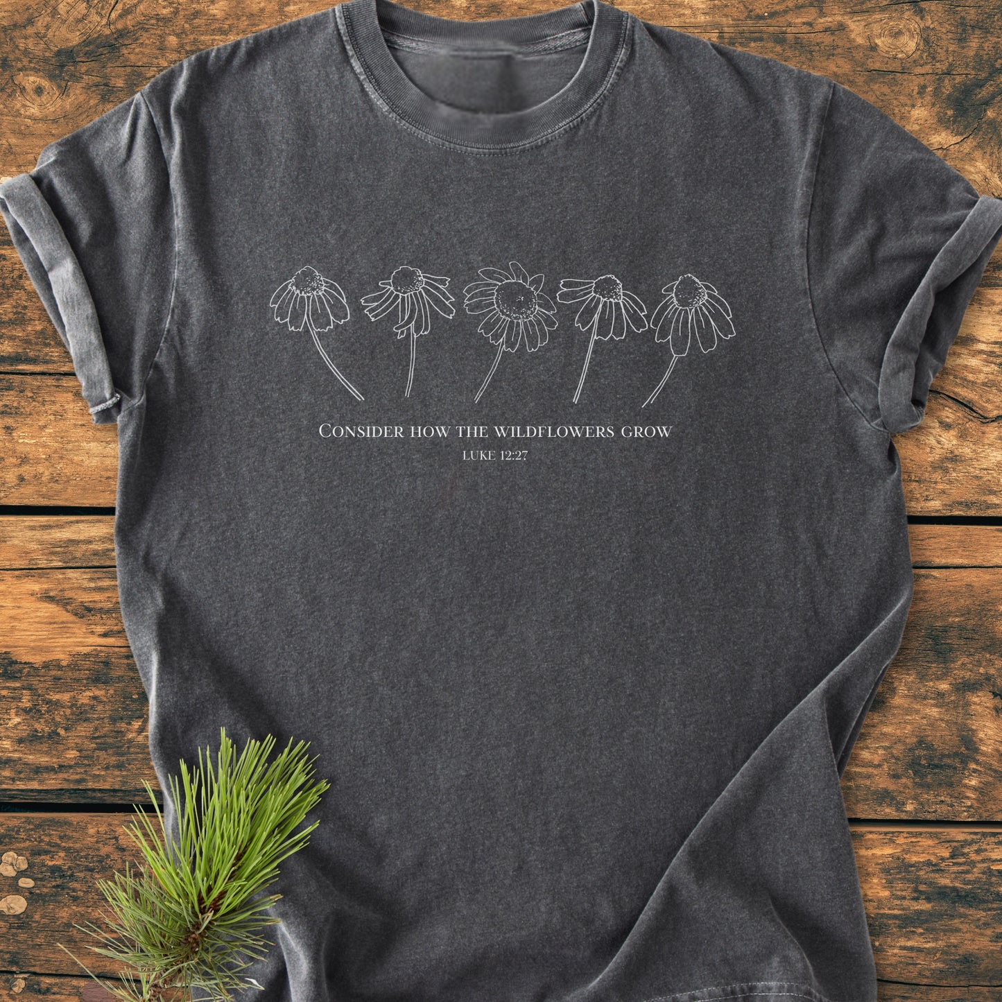 Consider How The Wildflowers Grow - Vintage Wash Tee featuring a white line drawing of wildflower heads and text, laid on a wooden surface with a pine sprig accentuating its rustic appeal.