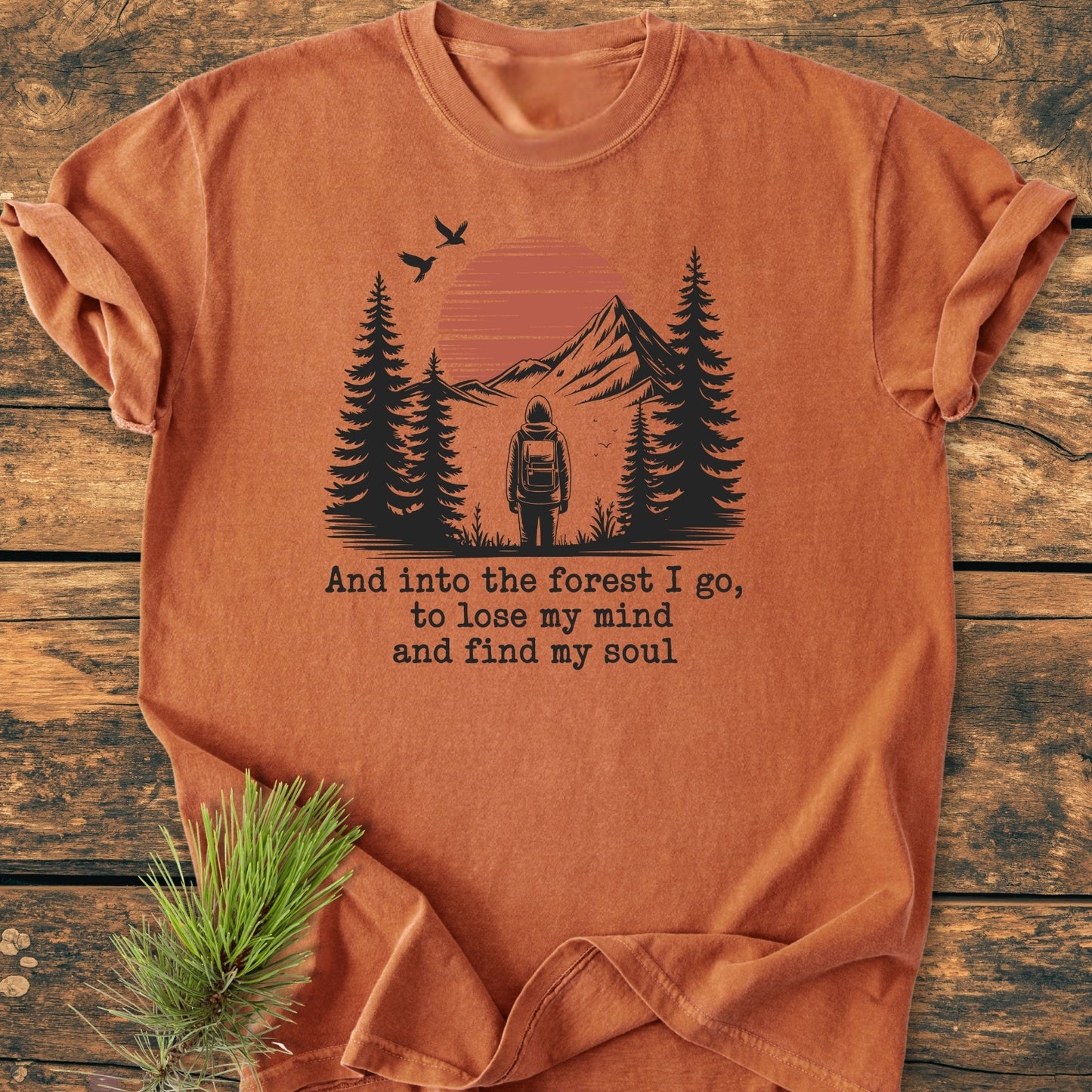 And Into The Forest I Go - Vintage Wash Tee: Rust-orange cotton t-shirt featuring a backpacker graphic amid pine trees and mountains, with typewriter-style forest quote, laid on rustic wood beside pine sprig.