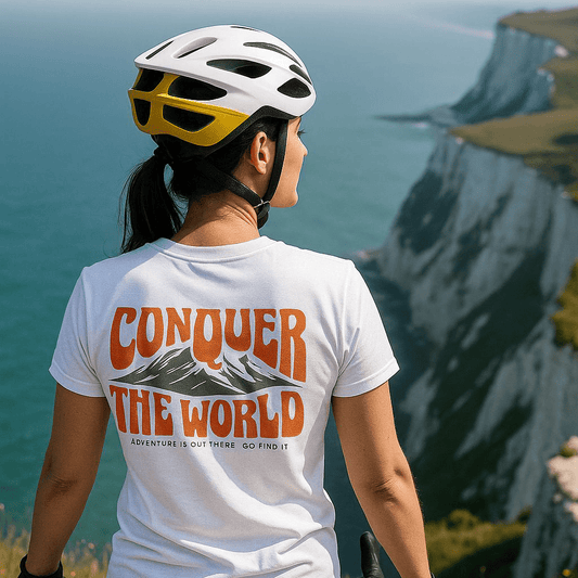 Person on cliff wearing Conquer The World T-shirt, featuring mountain graphic and slogan, with bike helmet and handlebars visible, overlooking dramatic ocean cliffs.