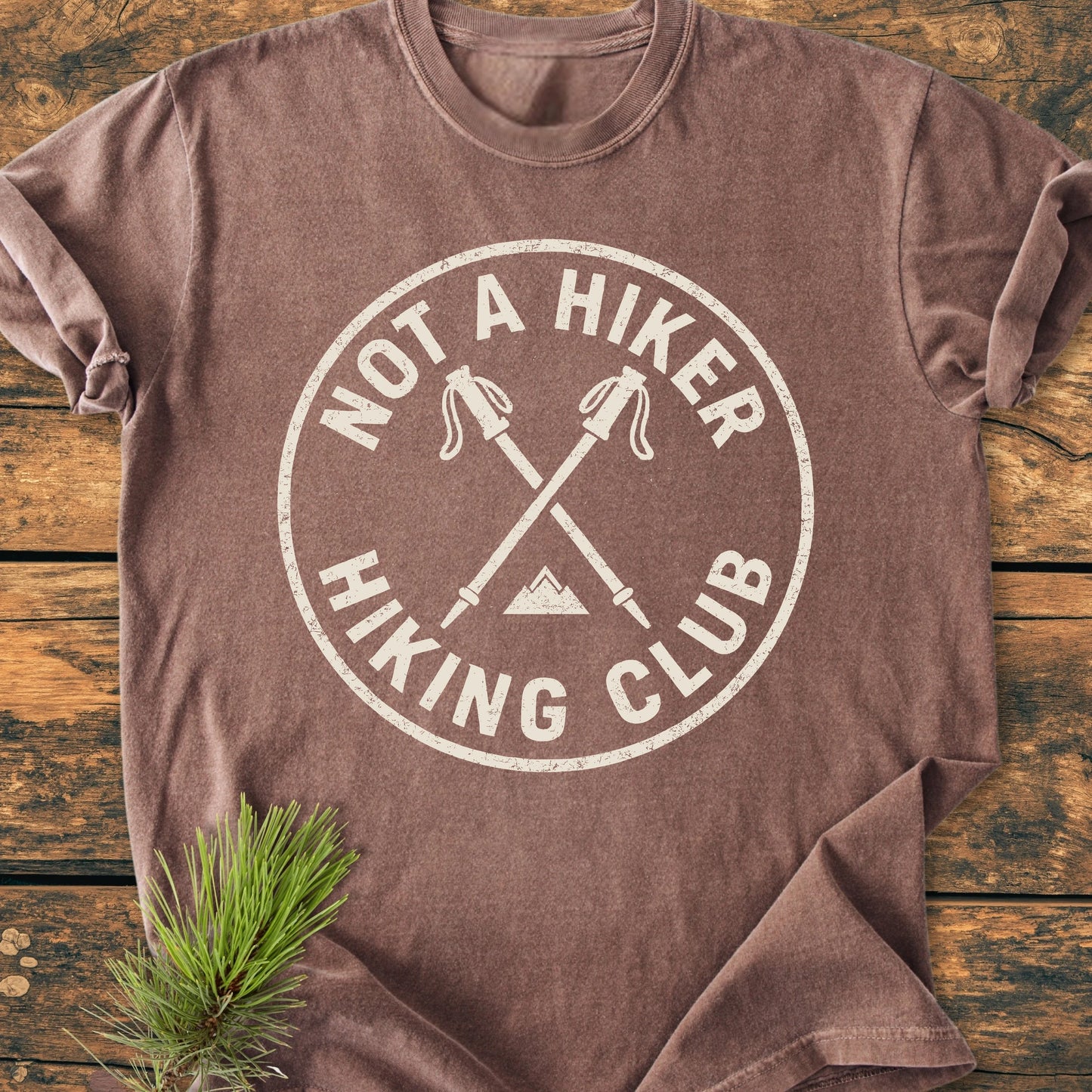 1426 Not A Hiker T-shirt with circular logo of crossed trekking poles and mountain graphic, laid flat on wood. Soft, vintage wash, unisex fit, perfect for outdoor enthusiasts seeking comfort and style.