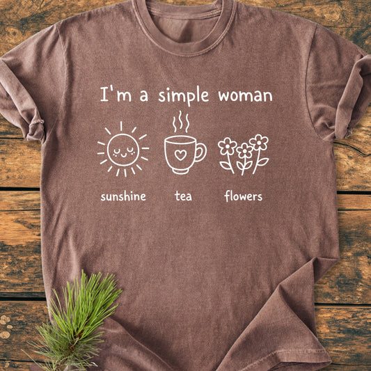 Brown 1408 I'm A Simple Woman T-shirt features white graphics with sunshine, tea, and flowers on a rustic wooden surface, with a pine sprig tucked under one edge.