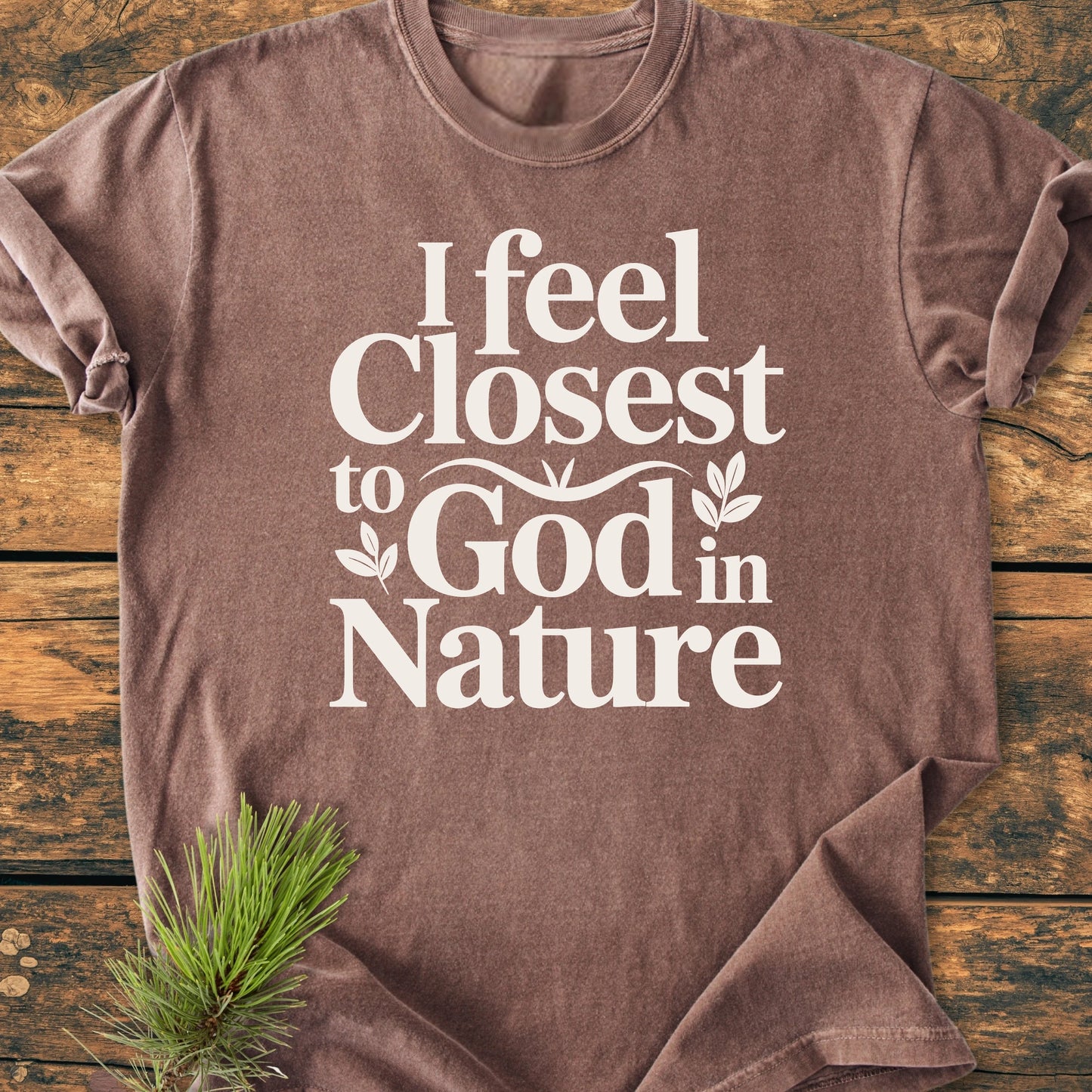 1427 Closest To God - Vintage Wash Tee with a white, bold graphic text and leaf illustrations on a soft brown shirt, laid on a wooden surface with a pine sprig underneath.