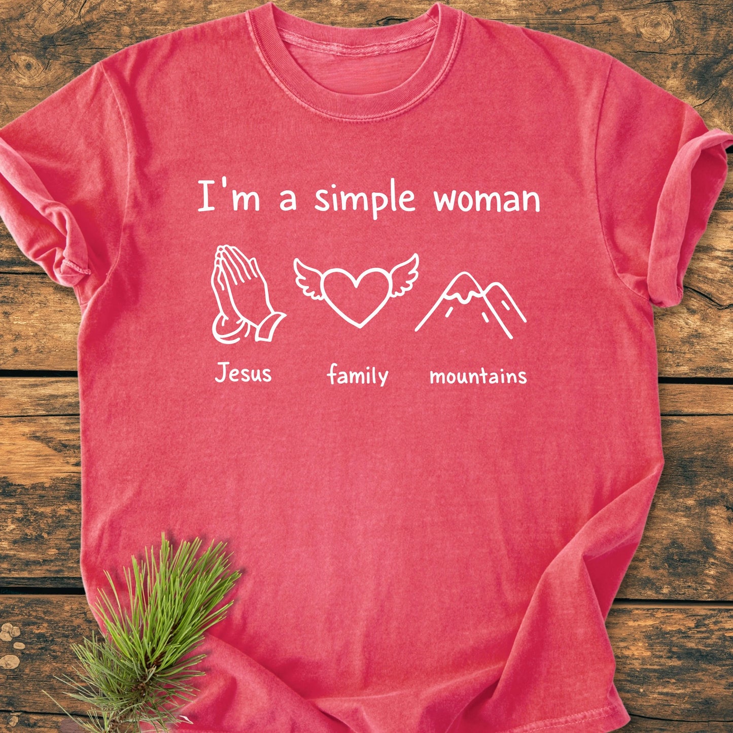 Heather-red T-shirt with “I’m a Simple Woman” and icons of praying hands, heart with wings, and mountains. Laid on wood with pine accent, embodying a vintage, relaxed lifestyle.