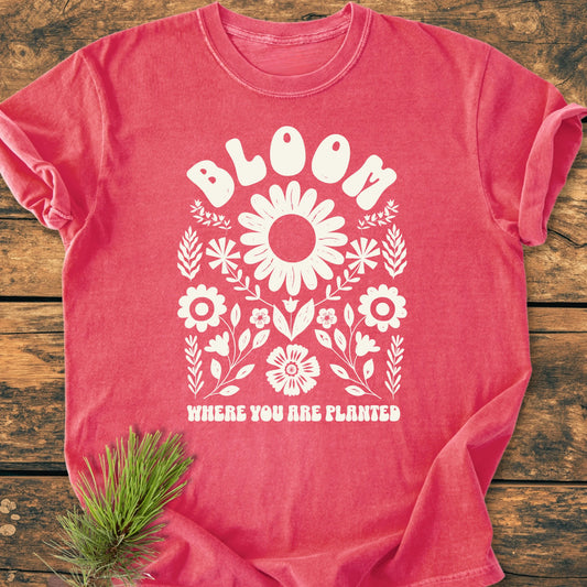 Coral-red cotton T-shirt with 1415 Bloom Where You Are Planted design featuring a large white daisy and floral motif, lying flat on a wooden surface with a pine twig beside it.