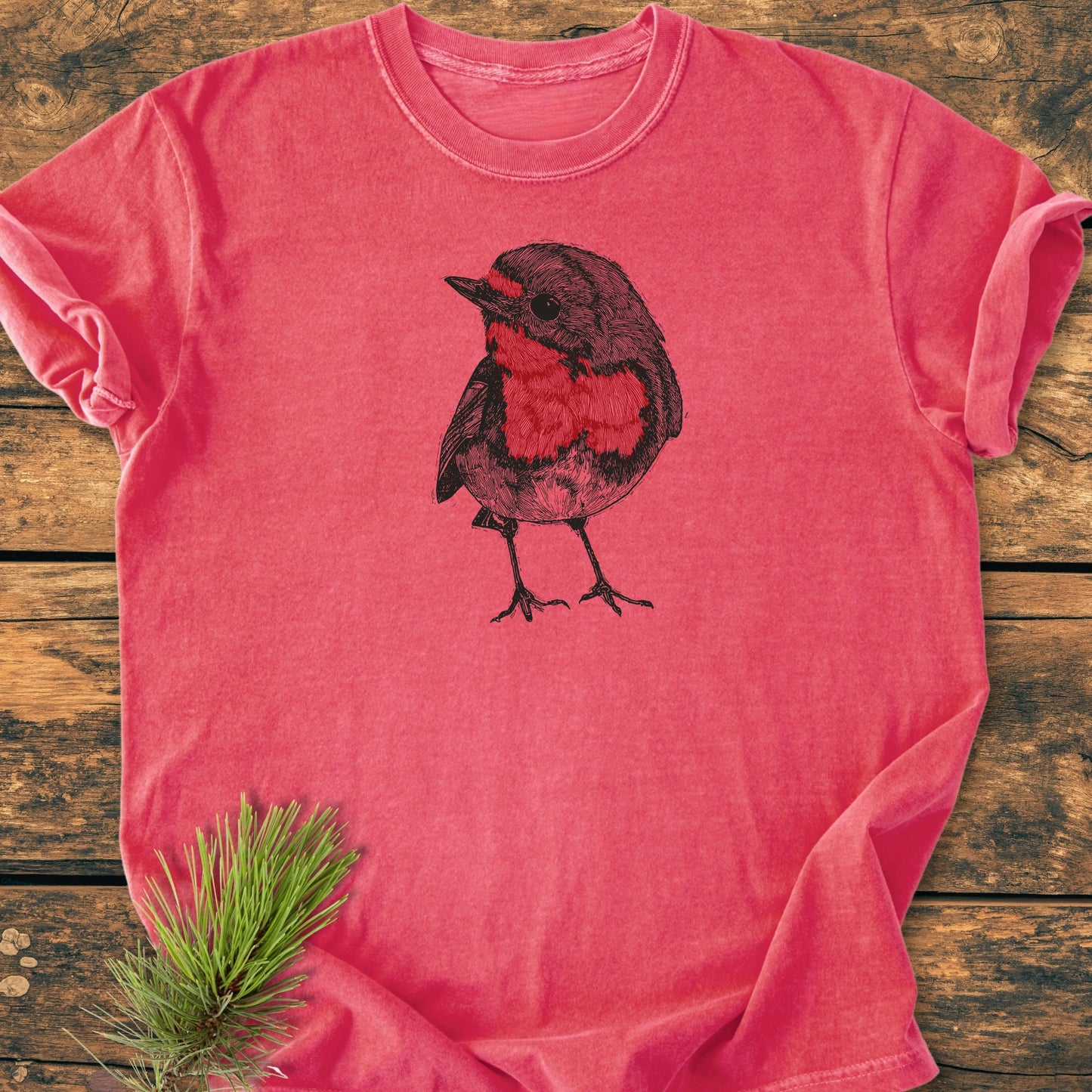 Cute Robin T-shirt laid flat on wood, features a hand-drawn robin illustration. Soft, vintage-wash fabric with a relaxed fit ideal for casual, everyday wear. Accented by a pine branch nearby.