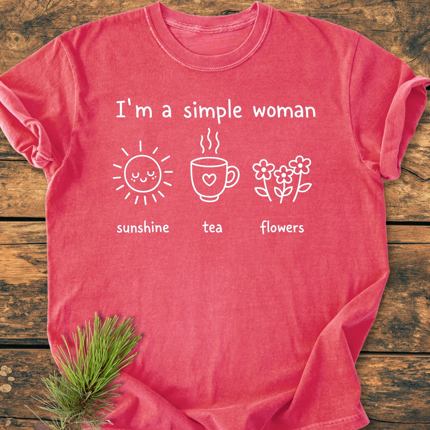 1408 I'm A Simple Woman T-shirt, showcasing a red-pink hue, I'm a simple woman text, icons of sunshine, tea, and flowers, styled with a pine sprig on a wooden surface.