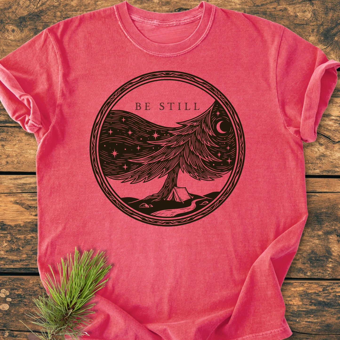 Pink-red 1411 Be Still T-shirt featuring a tree with starry sky design, crescent moon, tent, and path. Laid on wood with pine sprig, showcasing Comfort Colors' garment-dyed, ring-spun cotton fabric.