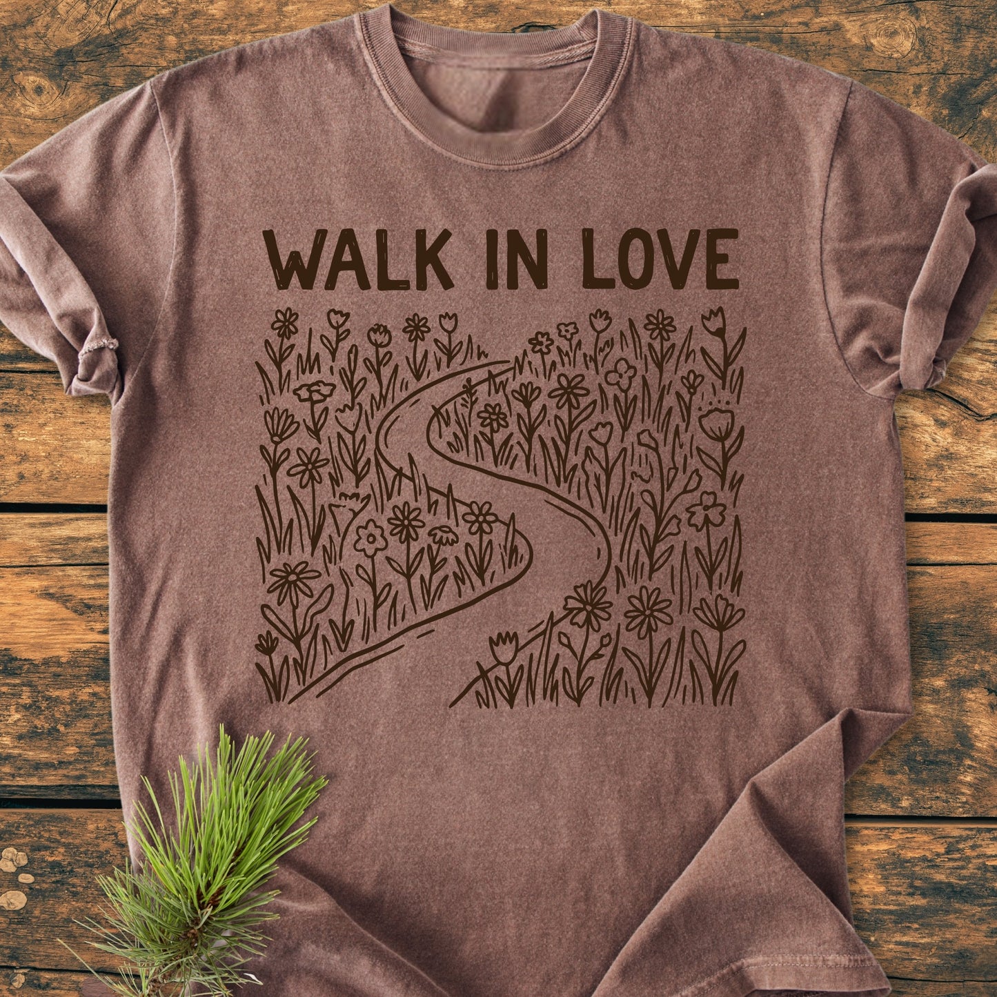 Walk In Love - Vintage Wash Tee: Heather-brown T-shirt with WALK IN LOVE text and meadow graphic on chest, laid flat on wood surface. Pine sprig accentuates the cozy, relaxed design.