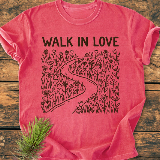Walk In Love - Vintage Wash Tee features a heather-red design with a black line-drawing of a path through flowers, bold WALK IN LOVE text, laid on a wooden surface with a pine branch accent.
