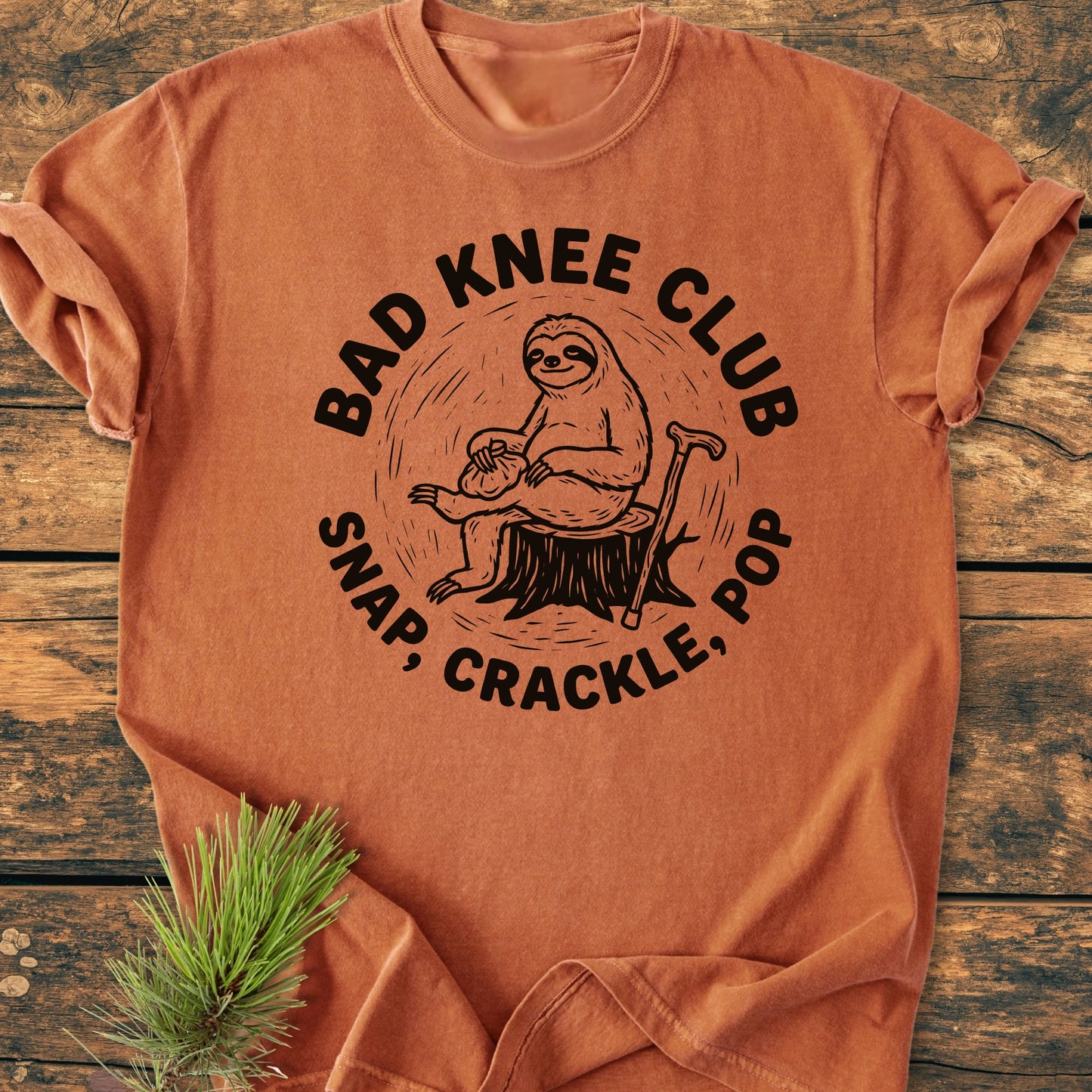 Cartoon sloth on a tree stump with a cane, captioned BAD KNEE CLUB and SNAP, CRACKLE, POP, printed on a vintage wash, soft, relaxed-fit T-shirt, ideal for everyday comfort.