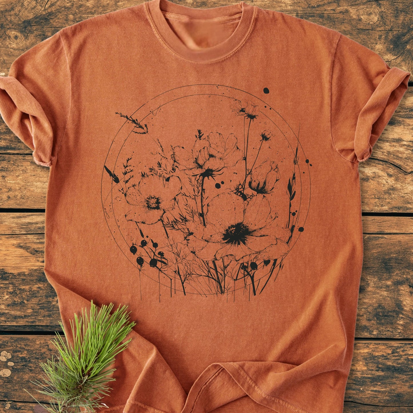 391 Wildflower Circle t-shirt: rust-orange crew-neck with a fine-line black botanical illustration on the front, displayed on wood. Includes poppy-like wildflowers within a circular frame, pine sprig accent.