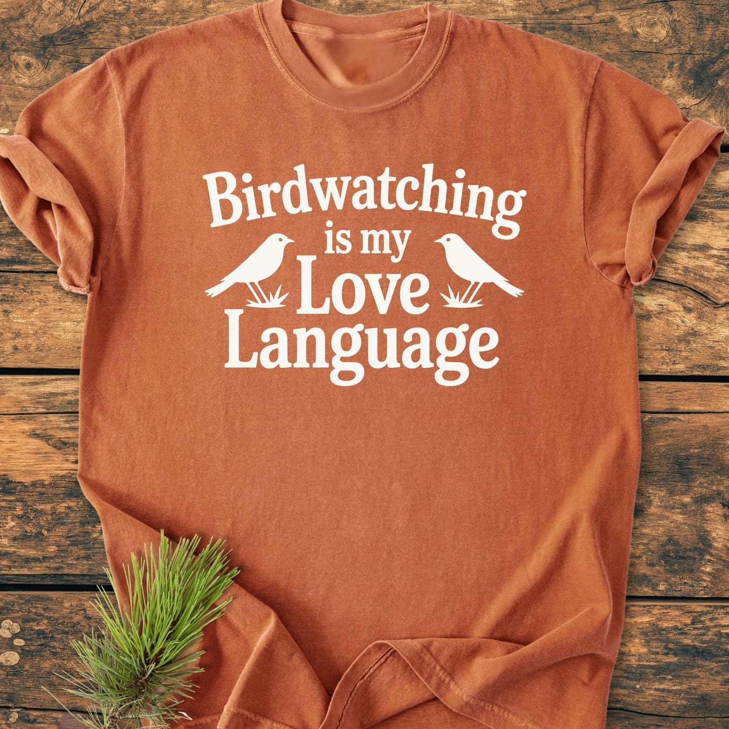 1428 Birdwatching Love Language T-shirt, russet-orange, with white bird graphic and text, laid flat on wooden surface. Pine sprig accentuates the vintage wash tee's outdoor-inspired design.