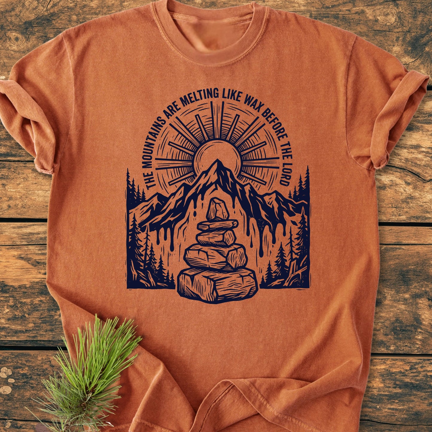 Rust-orange T-shirt titled 1414 Mountains Melting Like Wax, featuring a melting mountain range, sunburst, evergreen trees, and cairn, displayed on a wooden surface beside pine sprig.