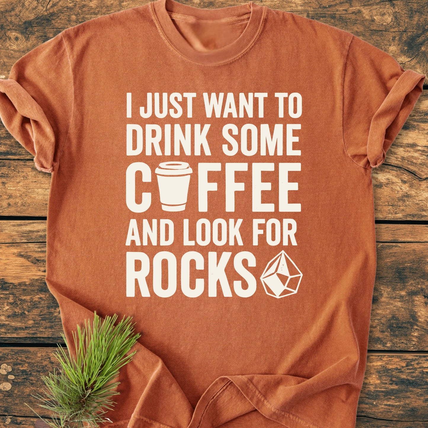 Rust-orange 1404 Drink Coffee And Look For Rocks Base T-shirt with bold slogan, coffee-cup icon, and gemstone graphic on front, laid flat on a wooden surface with pine sprigs.