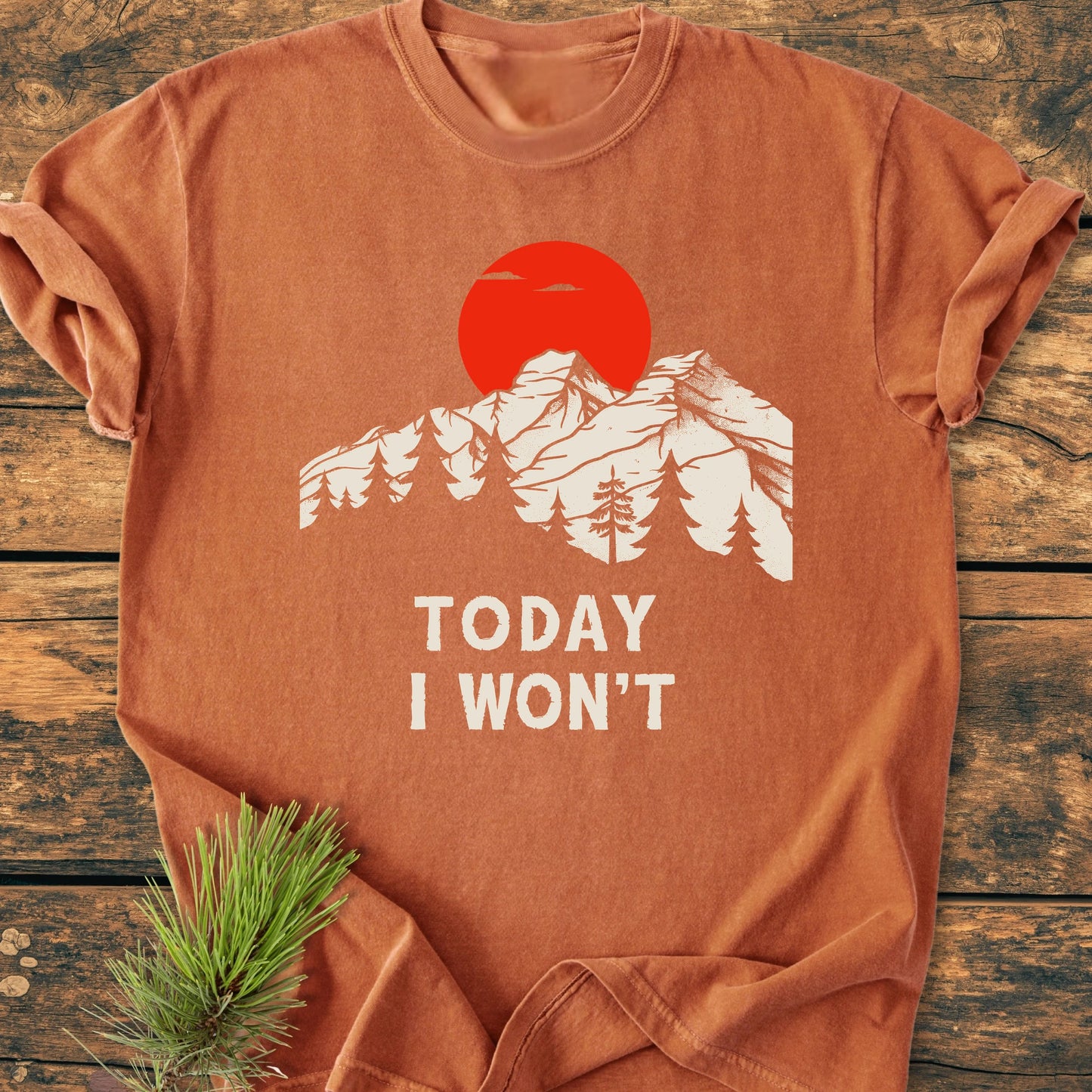 Rust-orange T-shirt with “TODAY I WON’T” text, featuring a white mountain and tree silhouette under a red sun. Laid on wood with pine sprig, reflecting Trails & Tees' vintage wash style.
