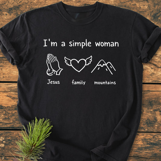 Black T-shirt with I'm A Simple Woman in white, featuring icons for Jesus, family, and mountains. Laid on wood with pine sprig, embodying a vintage, soft-washed, relaxed fit design.