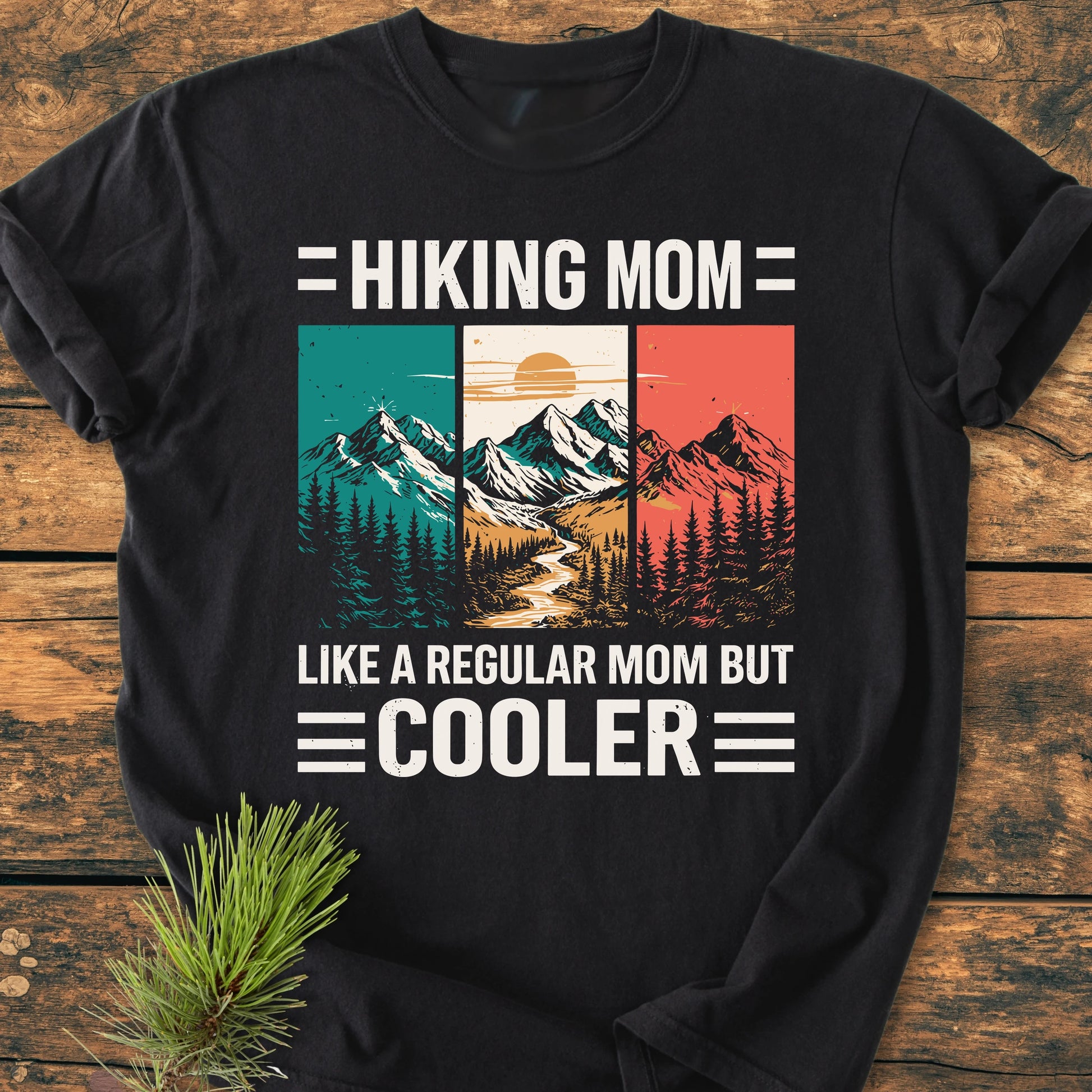 Hiking Mom - Vintage Wash Tee featuring a retro mountain scene with HIKING MOM and LIKE A REGULAR MOM BUT COOLER text, laid on wood with a pine sprig.
