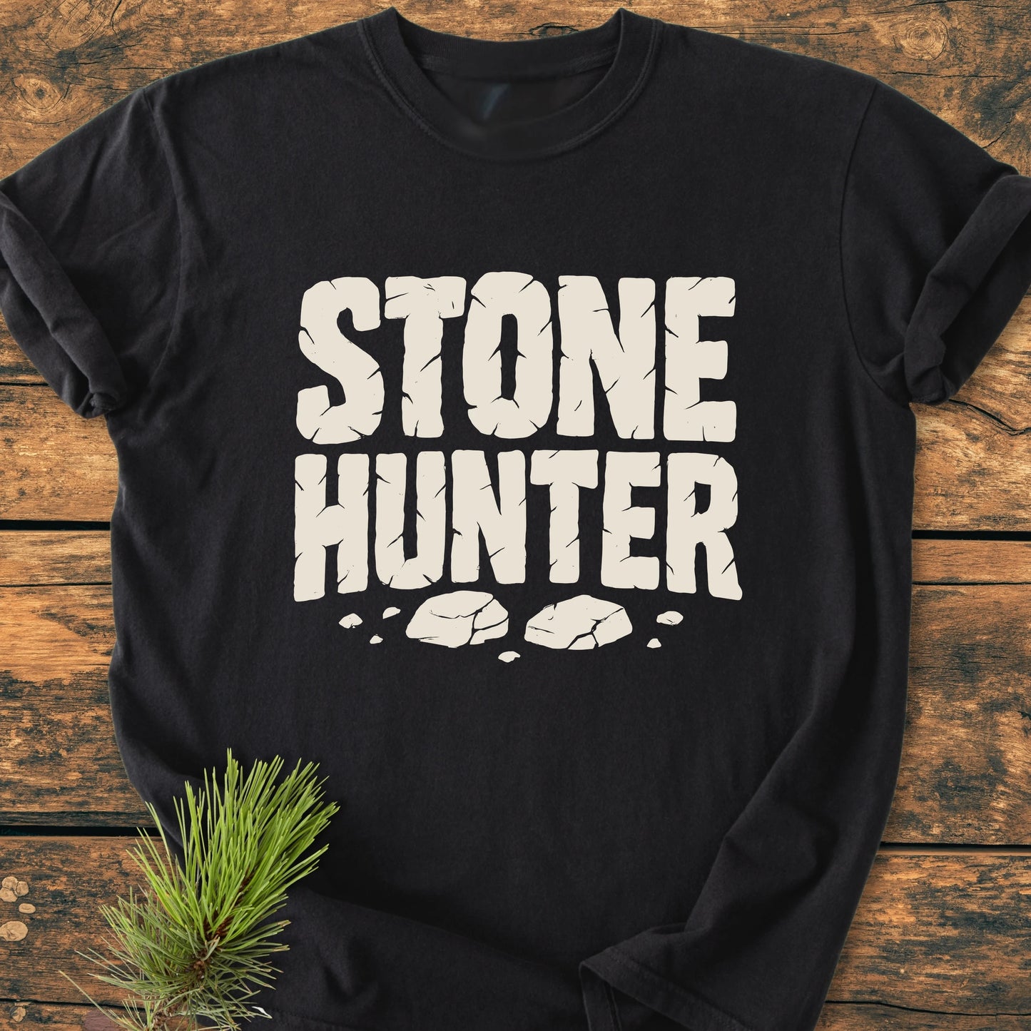 Black T-shirt titled 1425 Stone Hunter features cracked-stone–style white letters with rock illustrations. Laid on rustic wood, a pine branch adds natural detail, enhancing its vintage, outdoorsy appeal.