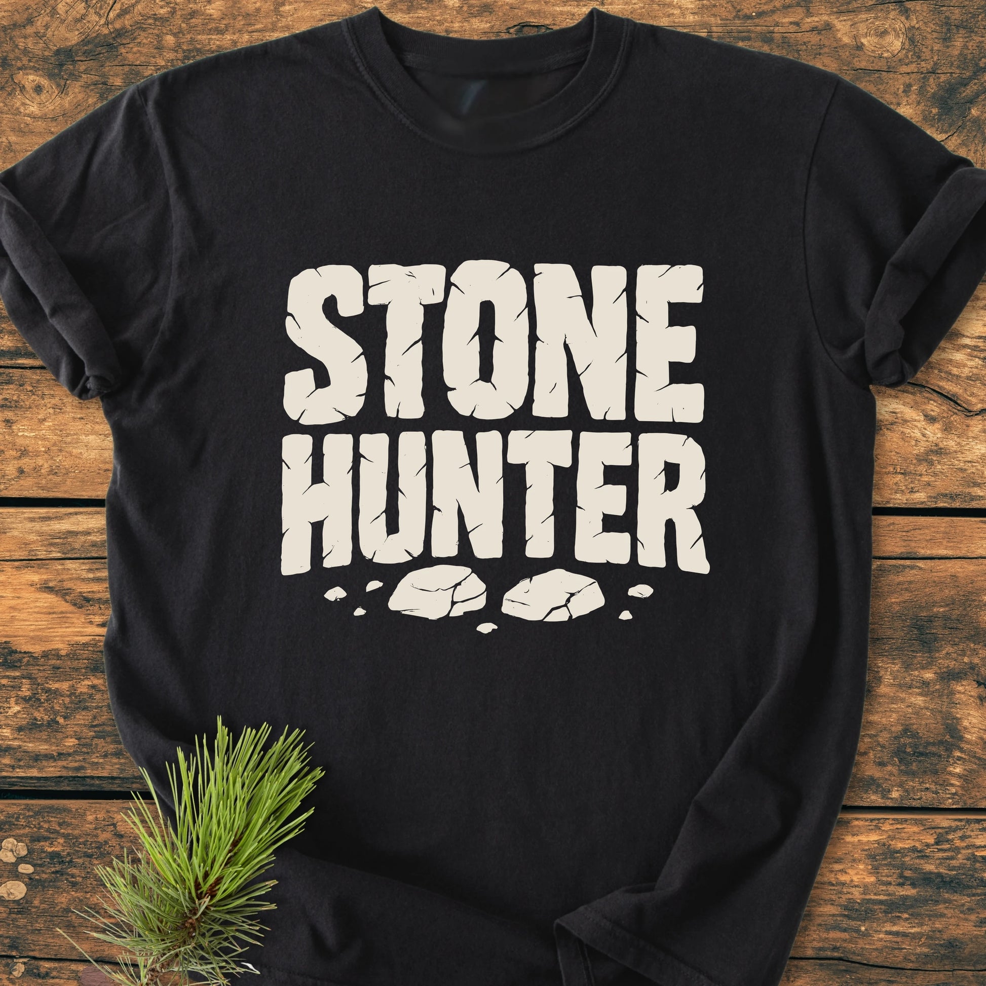 Black T-shirt titled 1425 Stone Hunter features cracked-stone–style white letters with rock illustrations. Laid on rustic wood, a pine branch adds natural detail, enhancing its vintage, outdoorsy appeal.