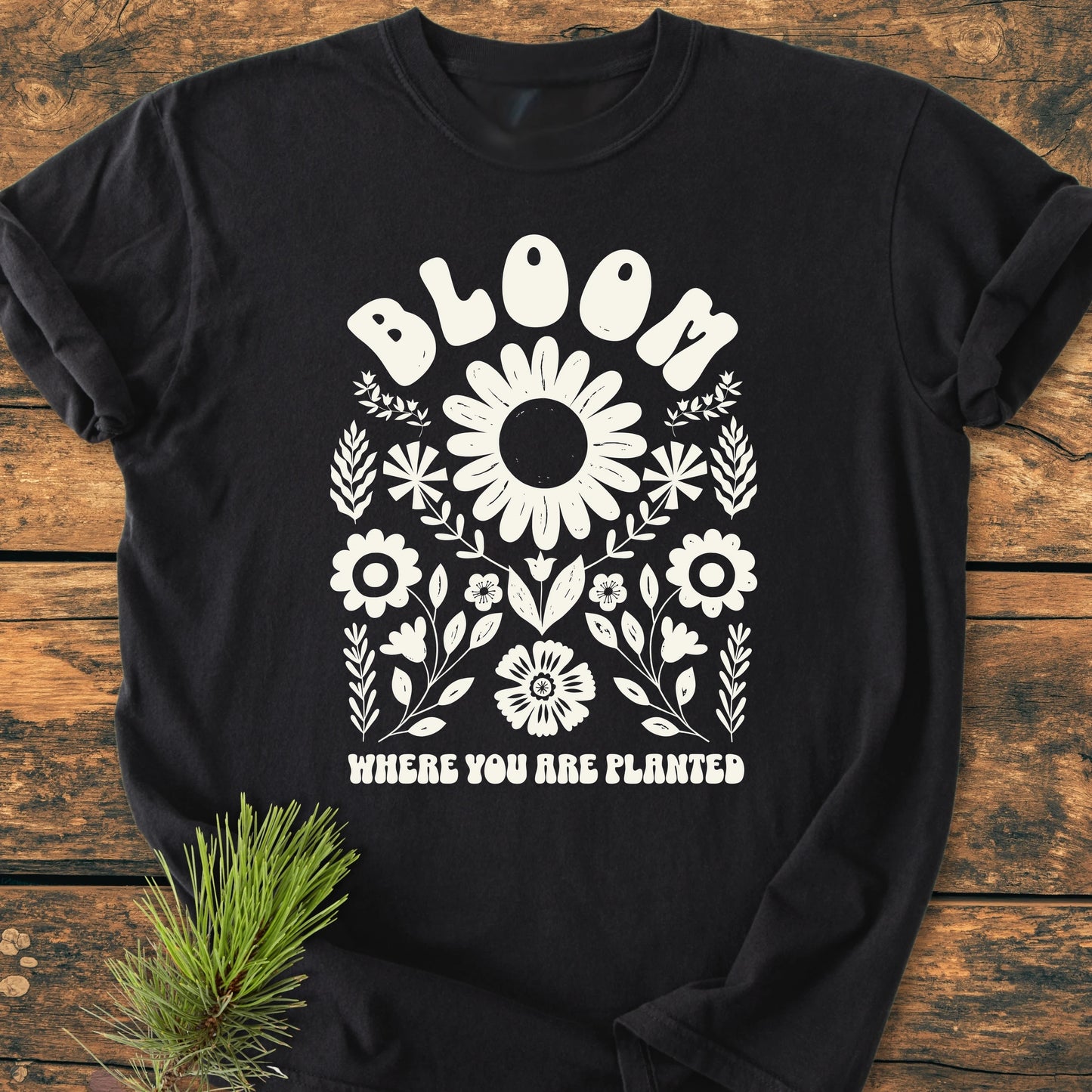 Black Bloom Where You Are Planted T-shirt featuring a white floral graphic with daisies. Laid flat on wood, it embodies a vintage wash and relaxed fit, perfect for everyday wear.
