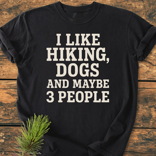 Black T-shirt, 1431 I Like Hiking And Dogs, featuring bold white text, laid flat on wood with a pine branch accent. Soft vintage wash, unisex relaxed fit, perfect for outdoor enthusiasts.