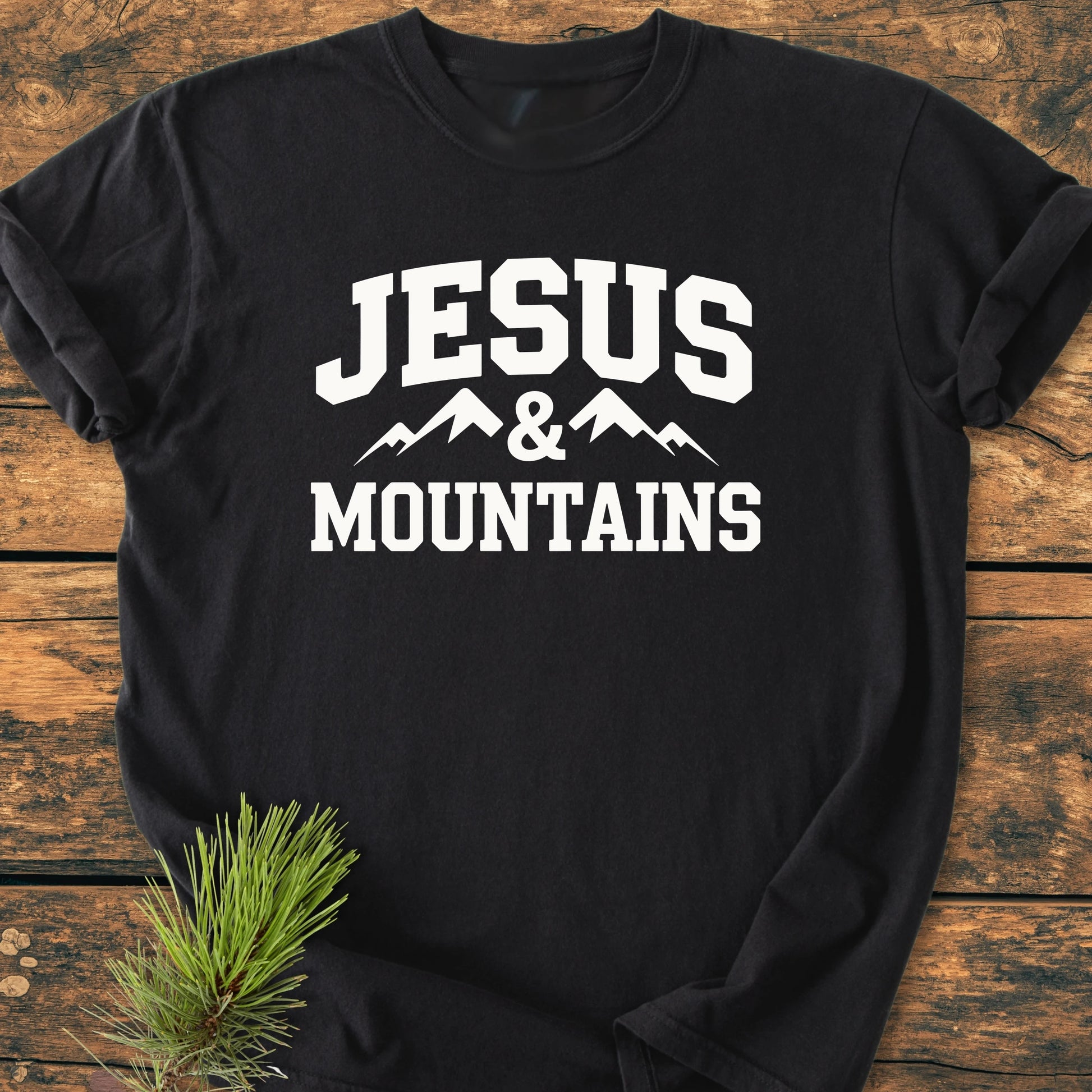 Black t-shirt with JESUS & MOUNTAINS in bold white letters, featuring mountain silhouette. Laid on rustic wood with pine needle accent, embodying outdoorsy and vintage feel.