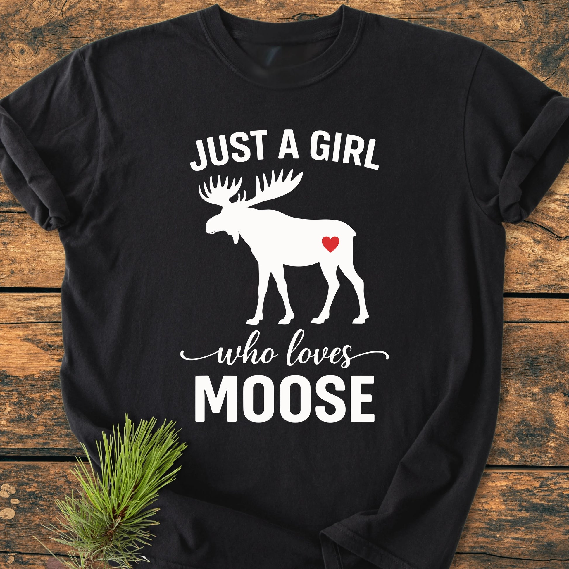 Black T-shirt titled 1437 Just A Girl Who Loves Moose, featuring a white moose silhouette with a red heart. Laid on wood surface with a pine sprig, highlighting its vintage, relaxed fit.