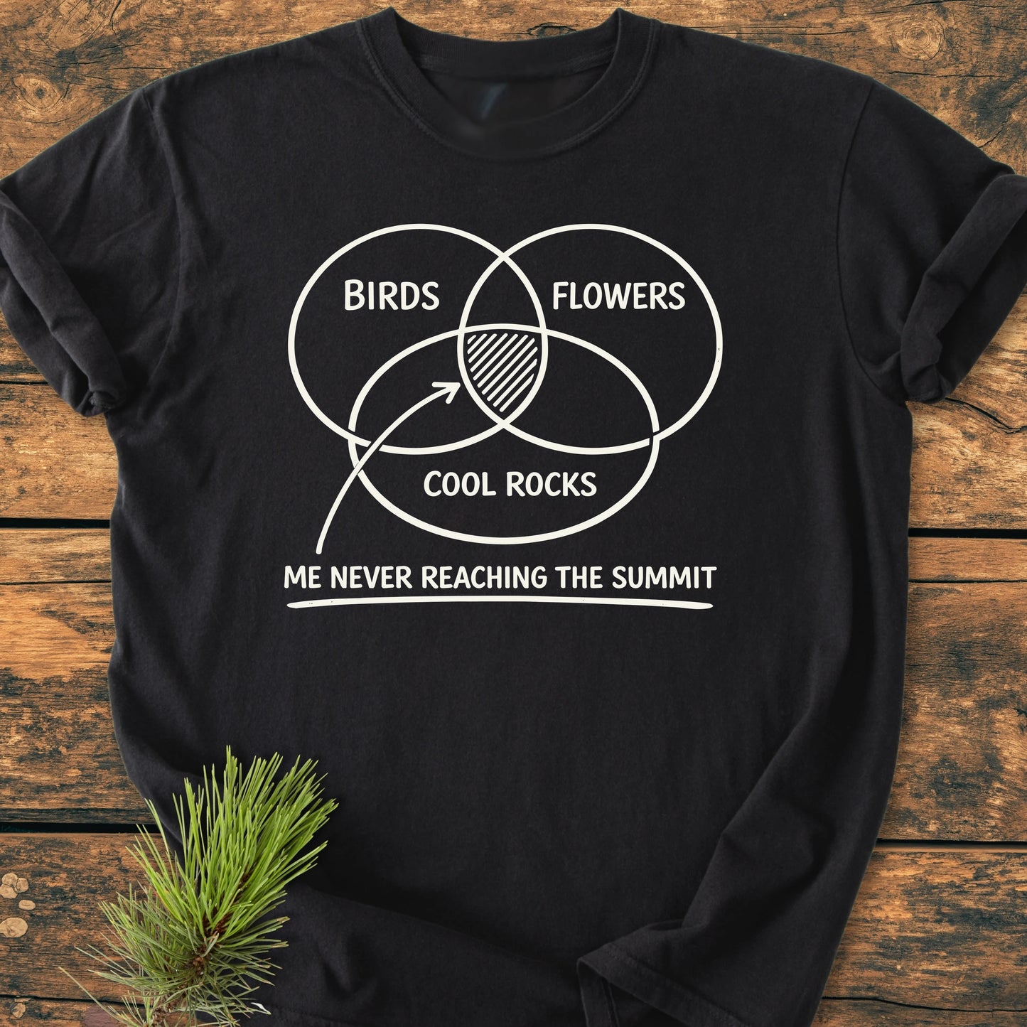 Black T-shirt titled 1416 Easily Distracted Hiker features a white Venn diagram of BIRDS, FLOWERS, COOL ROCKS with hatched overlap, captioned “ME NEVER REACHING THE SUMMIT.” Laid on wood surface.