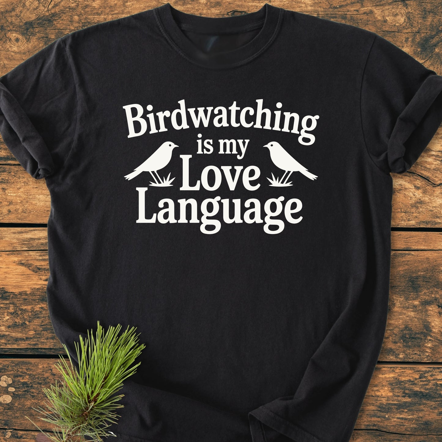 Black T-shirt titled 1428 Birdwatching Love Language features white text and bird silhouettes on the chest, laid flat with a pine sprig decoration, showcasing a soft, relaxed fit.