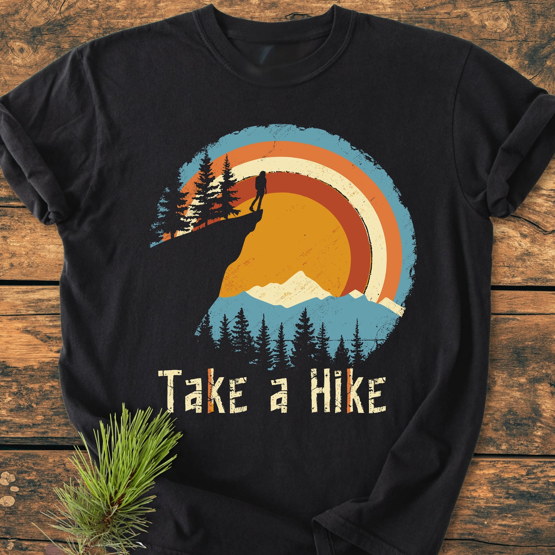 Take A Hike - Vintage Wash Tee: Black T-shirt with a vintage outdoor scene, featuring 'Take a Hike' text, hiker silhouette, and mountain backdrop. Laid on a wooden surface with a pine sprig.