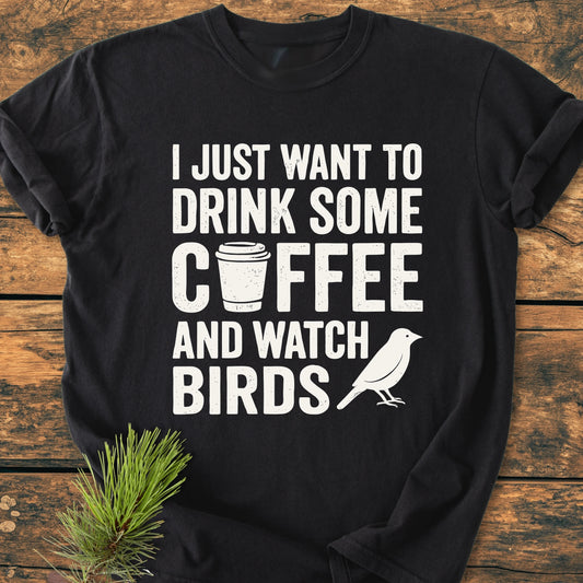 Black 1405 Drink Coffee And Watch Birds Base T-shirt with white screen print, featuring a coffee cup and bird silhouette, laid on a wooden surface with a pine sprig accent.