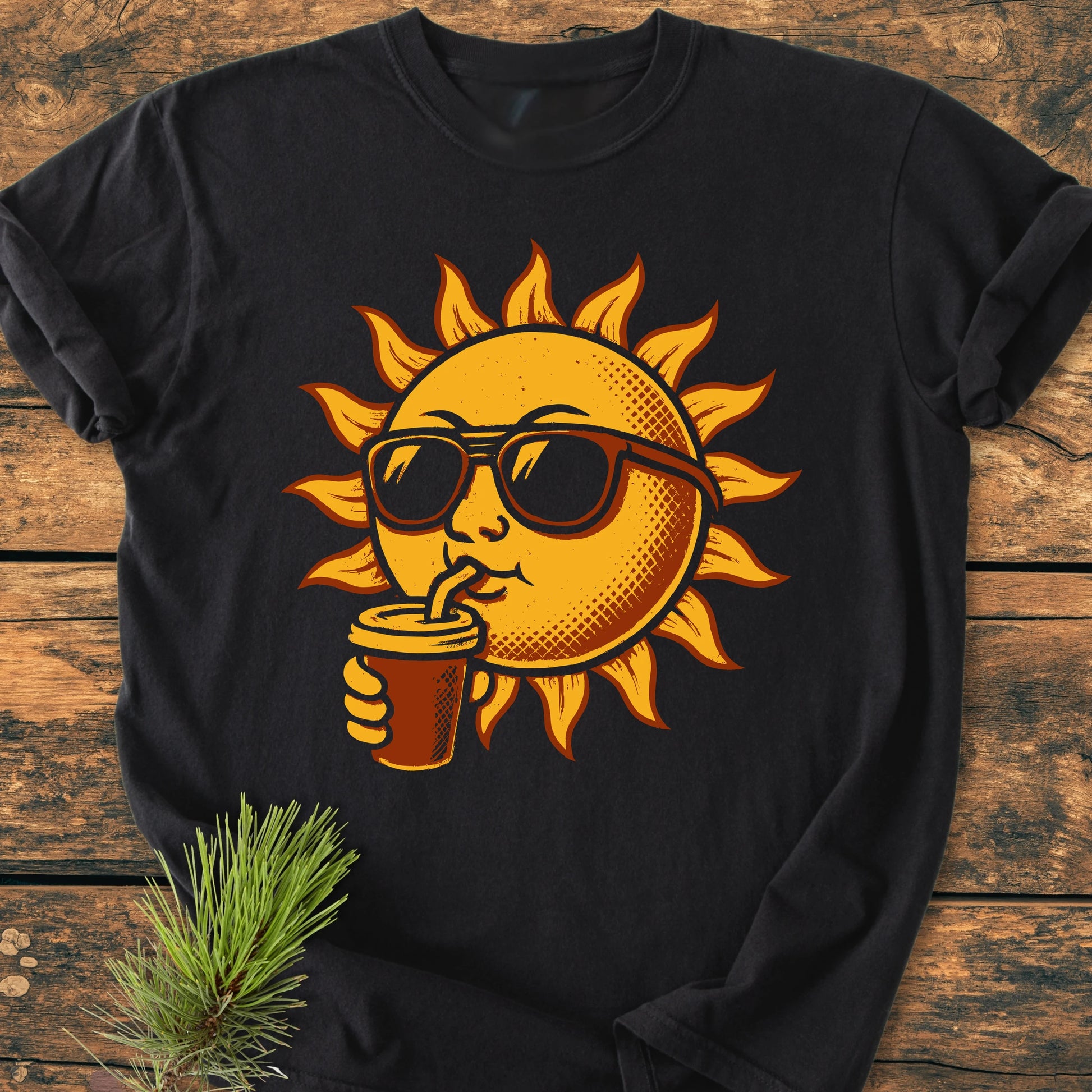 Black T-shirt 235 Sun With Coffee features a sun graphic with sunglasses sipping a drink; laid flat on wood with a pine sprig tucked under. Relaxed fit, garment-dyed, 100% cotton.