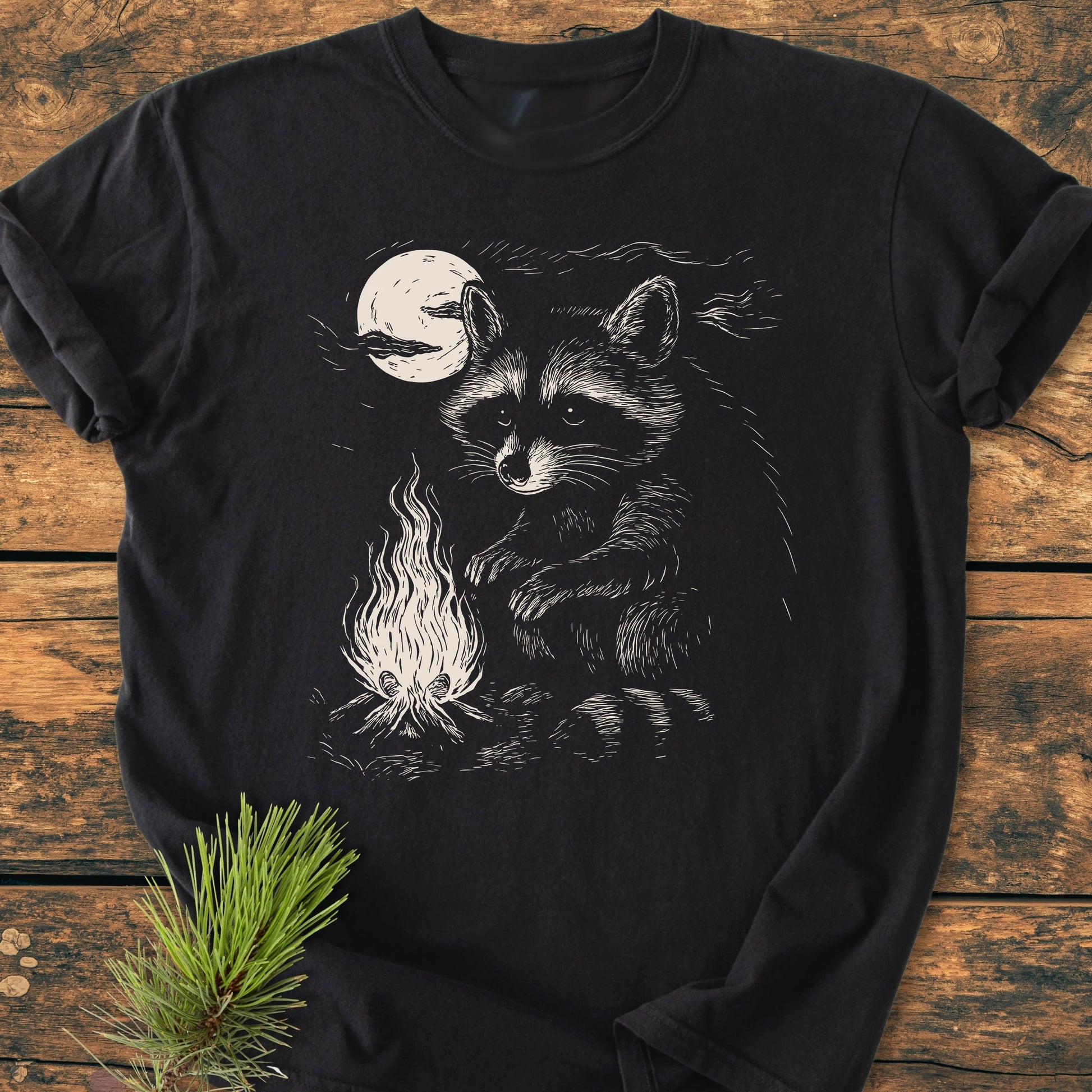 1409 Moonlit Campfire Raccoon Base T-shirt features a raccoon by a campfire under a moon, laid on a rustic surface with a pine sprig beside it, highlighting its cozy, durable design.