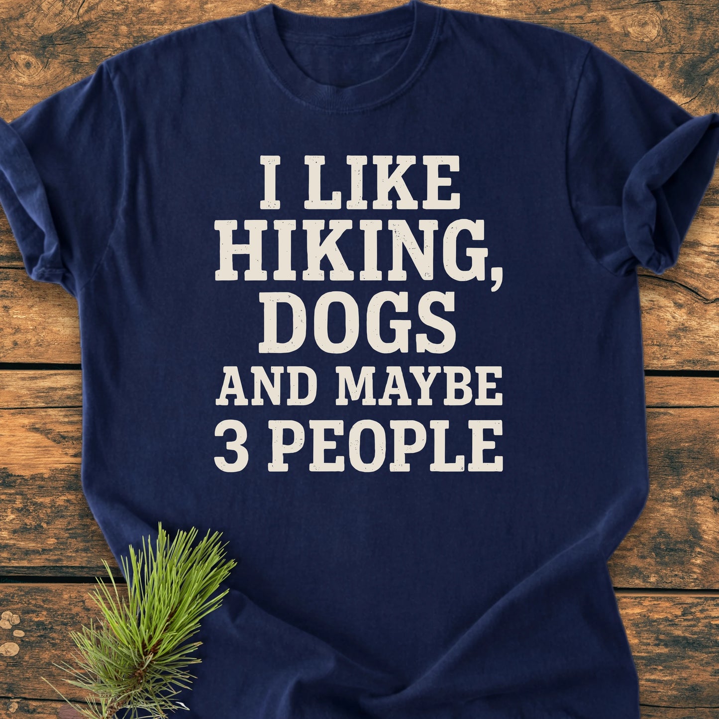 1431 I Like Hiking And Dogs navy tee with bold white text, laid on wood. Features pine needles accent, vintage wash, soft feel, and relaxed fit for everyday adventures.
