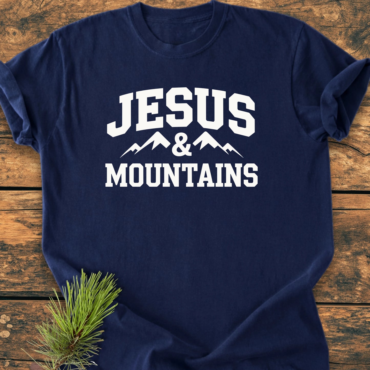 Navy-blue T-shirt with JESUS & MOUNTAINS in white, featuring a mountain graphic. Laid on wood, styled with a pine twig, showcasing a vintage wash, soft and relaxed fit.