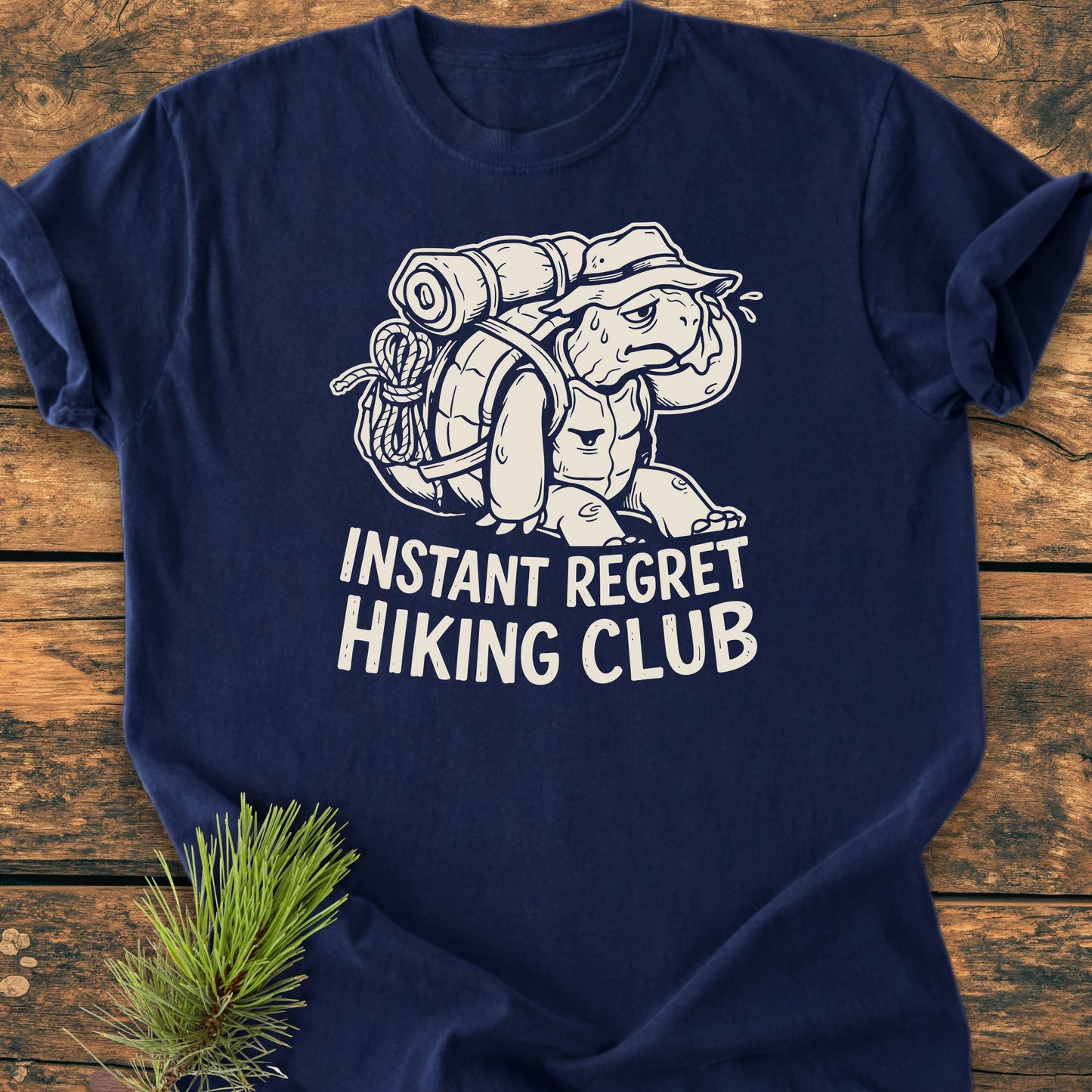 Navy-blue 1430 Instant Regret Hiking Club T-shirt with cartoon turtle in sun hat and hiking pack, slogan beneath. Laid flat with pine sprig accent, showcasing vintage wash and relaxed fit.