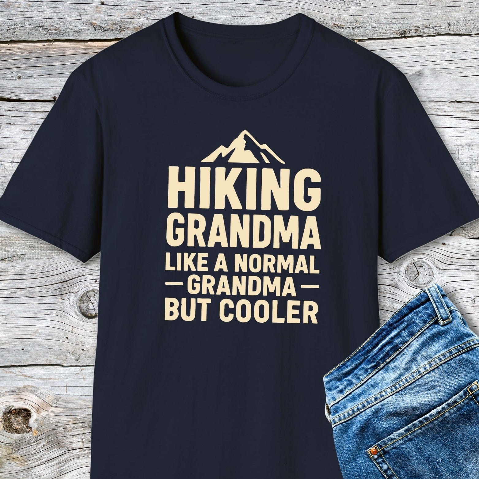 Hiking Grandma T-Shirt with mountain graphic and bold text, laid beside folded jeans on wooden planks, showcasing its adventure-ready design and comfortable fit.