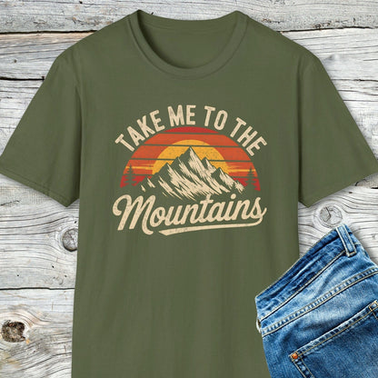 Take Me To The Mountains T-Shirt featuring retro sunset/mountain graphic, laid next to blue jeans; soft, lightweight cotton with unisex fit, ideal for adventures.