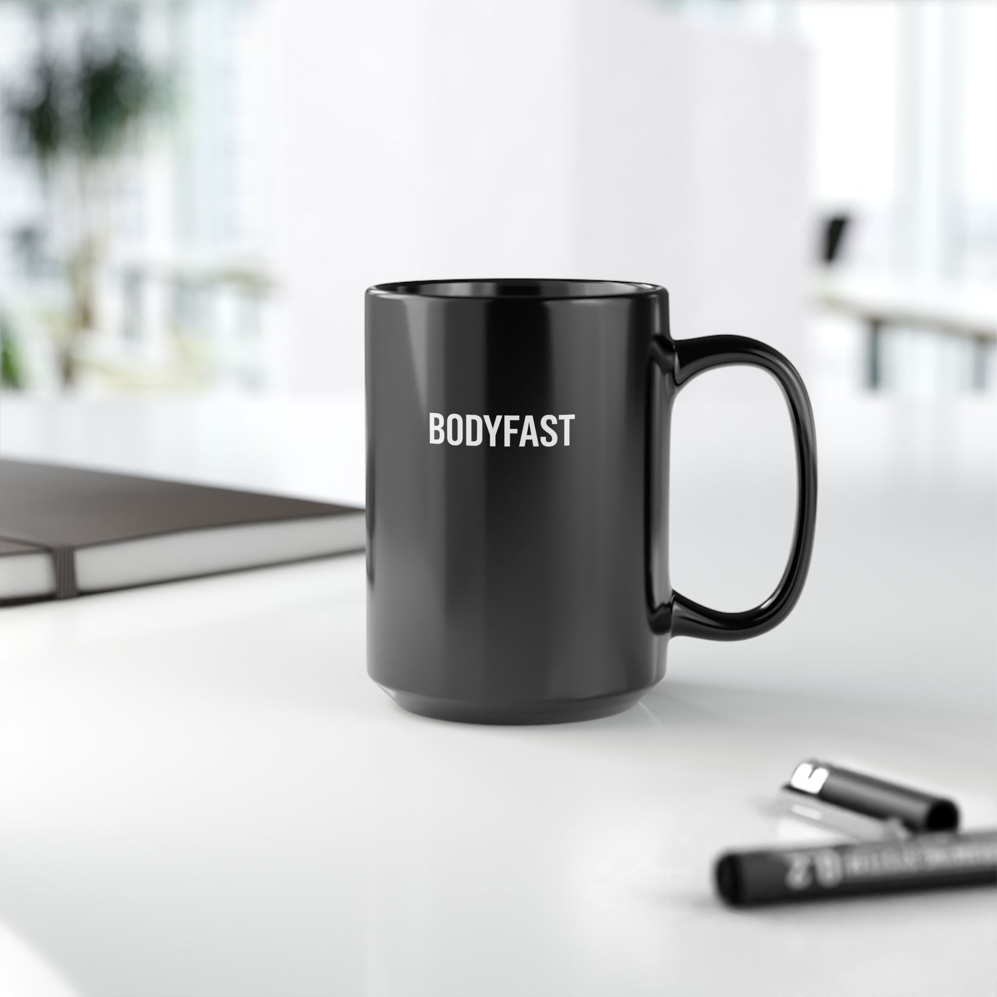 BODYFAST Ceramic Mug on white tabletop, featuring bold white text. Accompanied by a closed notebook and pens, with an office setting softly blurred in the background.