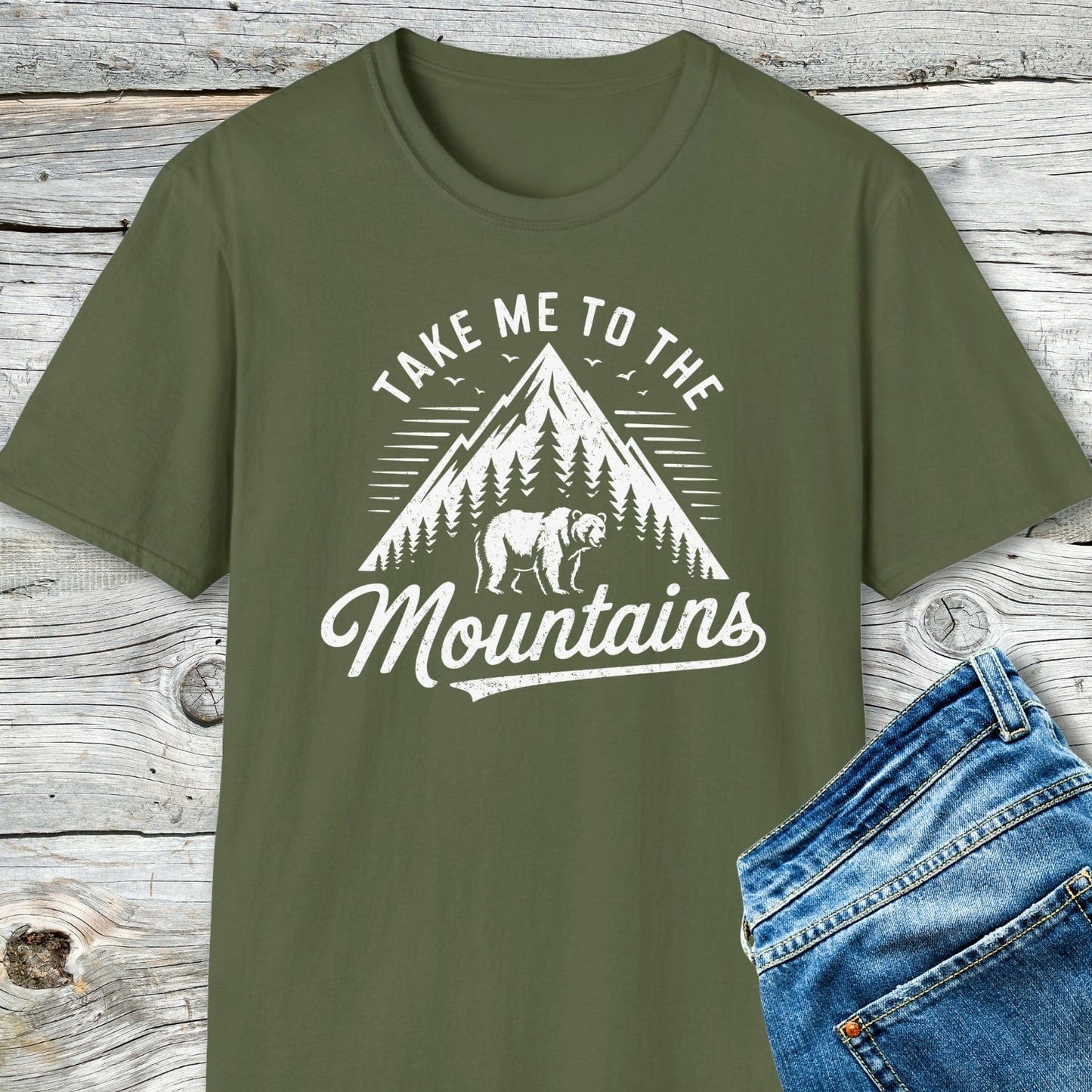 Take Me To The Mountains T-Shirt with mountain and bear motif, paired with blue jeans, showcased on a rustic wooden surface. Soft, lightweight cotton for adventure.