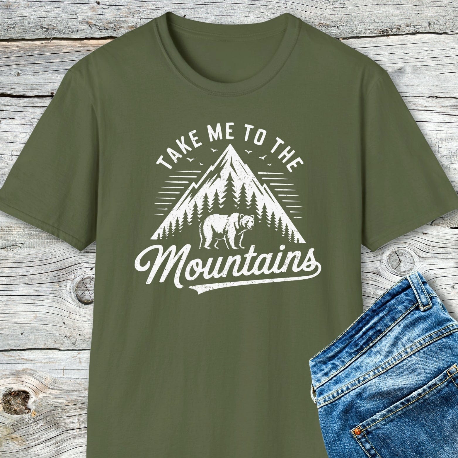 Take Me To The Mountains T-Shirt with mountain and bear motif, paired with blue jeans, showcased on a rustic wooden surface. Soft, lightweight cotton for adventure.